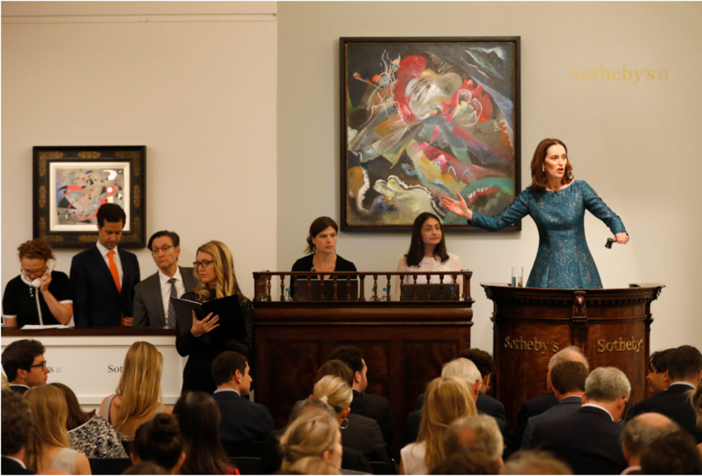 Photograph of a fine art auction in progress featuring Post-War and Contemporary paintings. A woman auctioneer wearing a blue dress stands at the podium wearing a blue dress while a crowd views the art on offer.