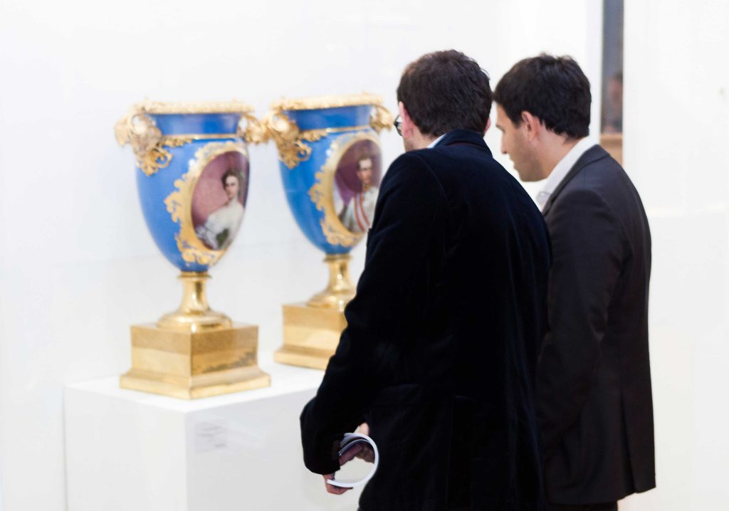 An image of two men in suits viewing two decorative classical vases featuring portrait paintings on display.