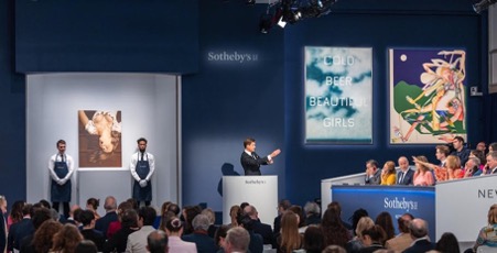 Portrait of Sotheby’s Senior International Specialist and Auctioneer Michael Macaulay during “The Now” evening auction, 2022. A large audience watches a man, the auctioneer, at a podium point to two paintings. They are sitting in Sotheby's auction room surrounded by dark blue walls.