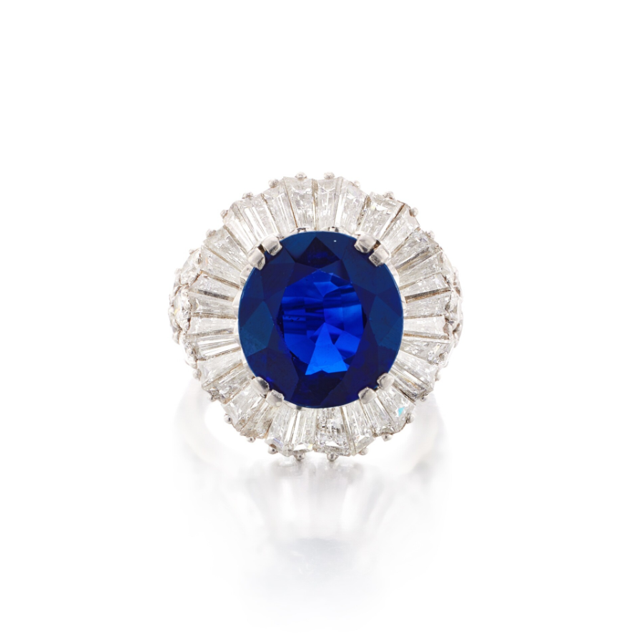A photograph of a ring of size 5, centering an oval-shaped sapphire, framed by tapered baguette diamonds, the shoulders further accented by round diamonds.