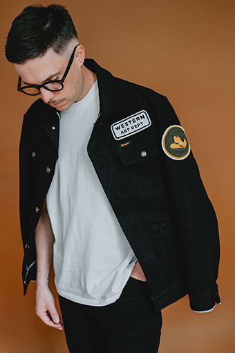 Photograph of artist Jeremy Booth in front of an orange background modeling his black jean jacket embroidered with, "Western Art Department."