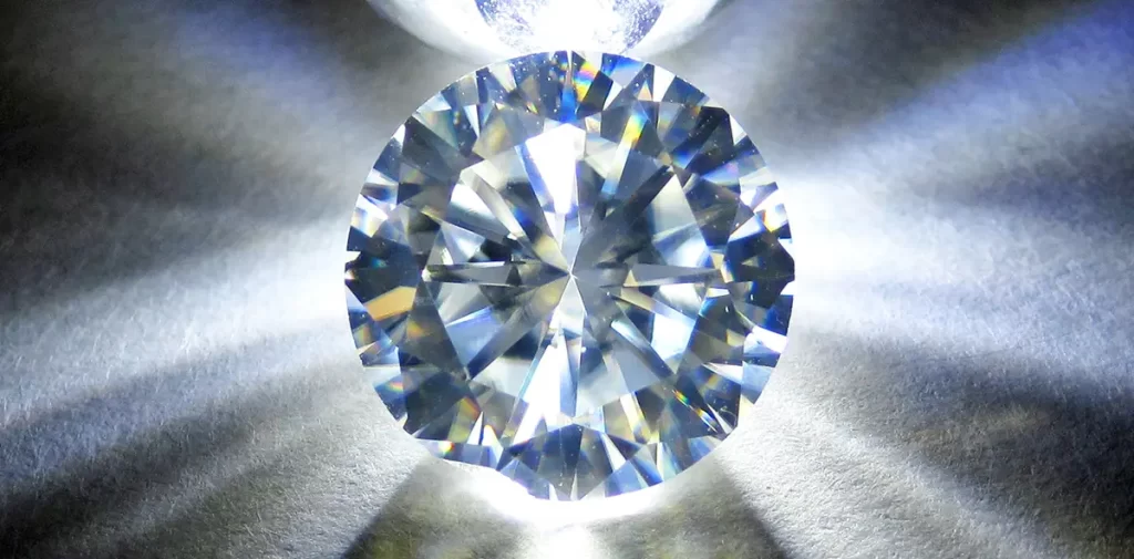 A macro photograph of a round, faceted, white moissanite, an imitation diamond similar to cubic zirconias, with a bright light behind the stone.