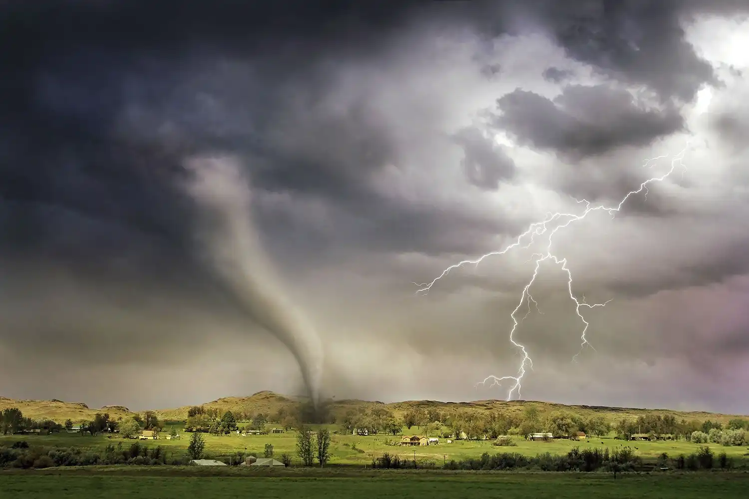 A photograph of a green landscape featuring both a tornado and lightening in the sky above.