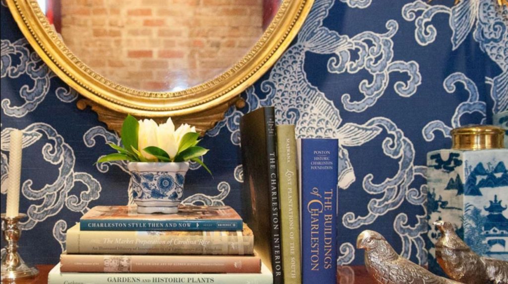 Close-up photo of a tabletop with books, decorative arts, and a gold mirror with a patterned blue wallpaper behind