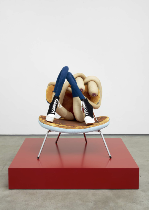 This image by Sarah Lucas depicts a low chair on a red stage in a white room. On the chair is two legs in Converse Chuck Taylor inspired high heels leading to a mass of legs in a knot-like form with parts that look like the female body.