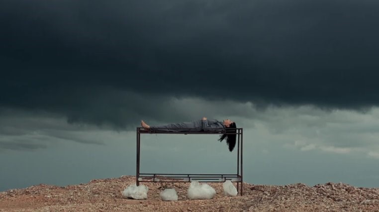 This image is of Marina Abramovic laying on top of a metal structure on a mountain top. There are grey skies and thunder clouds in the background.