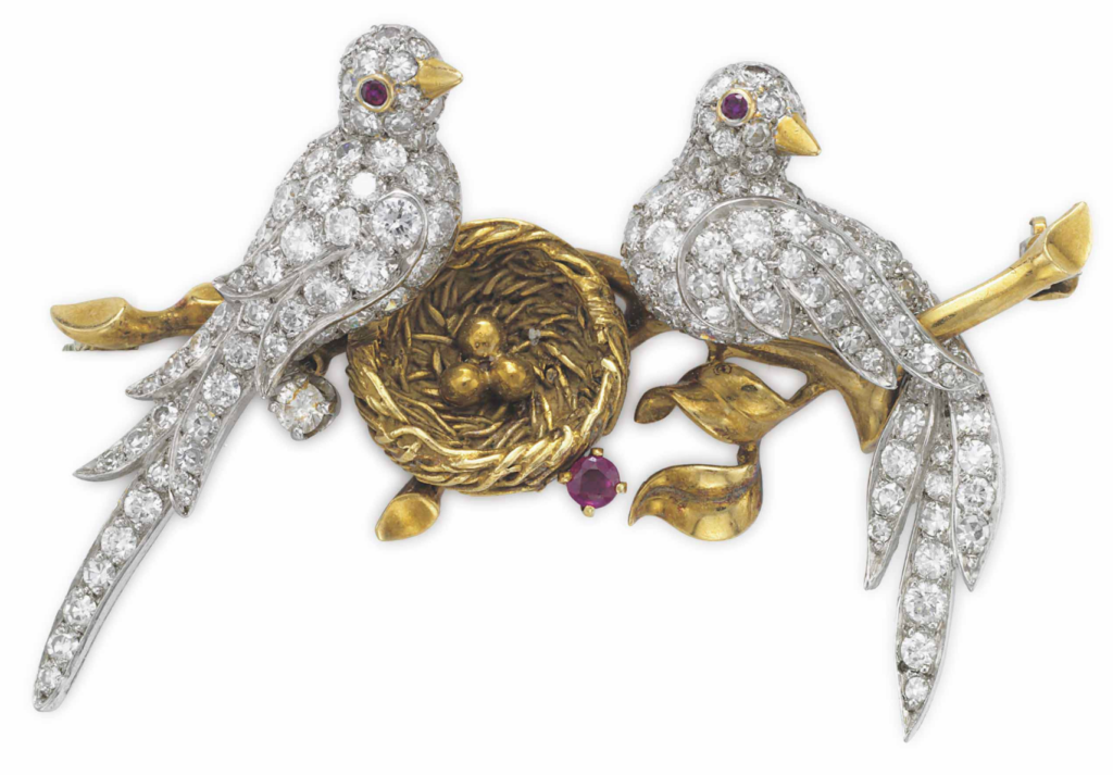 A gold, white diamond, and red ruby elaborate brooch shaped like two love birds sitting on golden branches beside a golden nest with eggs by luxury jewelry brand Van Cleef & Arpels