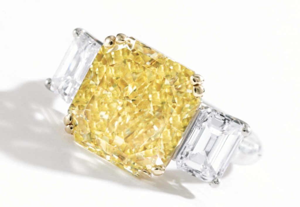 A jeweled ring, set with a large yellow square diamond with two rectangular white diamonds on either side, set against a white studio background.